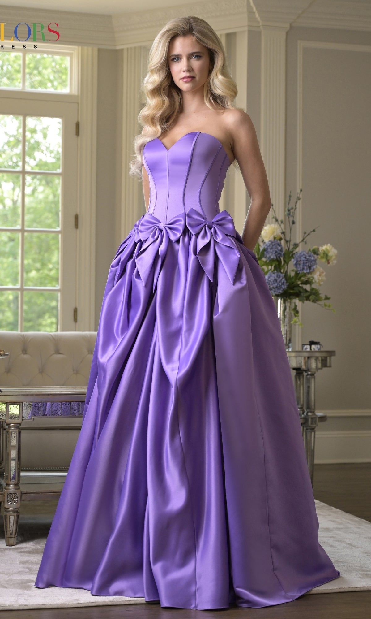 A woman stands indoors wearing the Bow-Front Long Prom Ball Gown by Colors Dress 3739—a floor-length lavender A-line prom dress with a fitted bodice, voluminous skirt, and strapless sweetheart neckline.